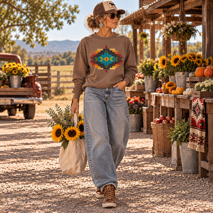 A woman stands on a gravel farm road wearing a brown long-sleeve crewneck sweatshirt with a colorful geometric pattern on the chest, paired with light-wash wide-leg jeans and brown lace-up boots. She is holding a canvas tote bag filled with sunflowers, with a rustic farm stand, flowers, and a vintage pickup truck in the background.