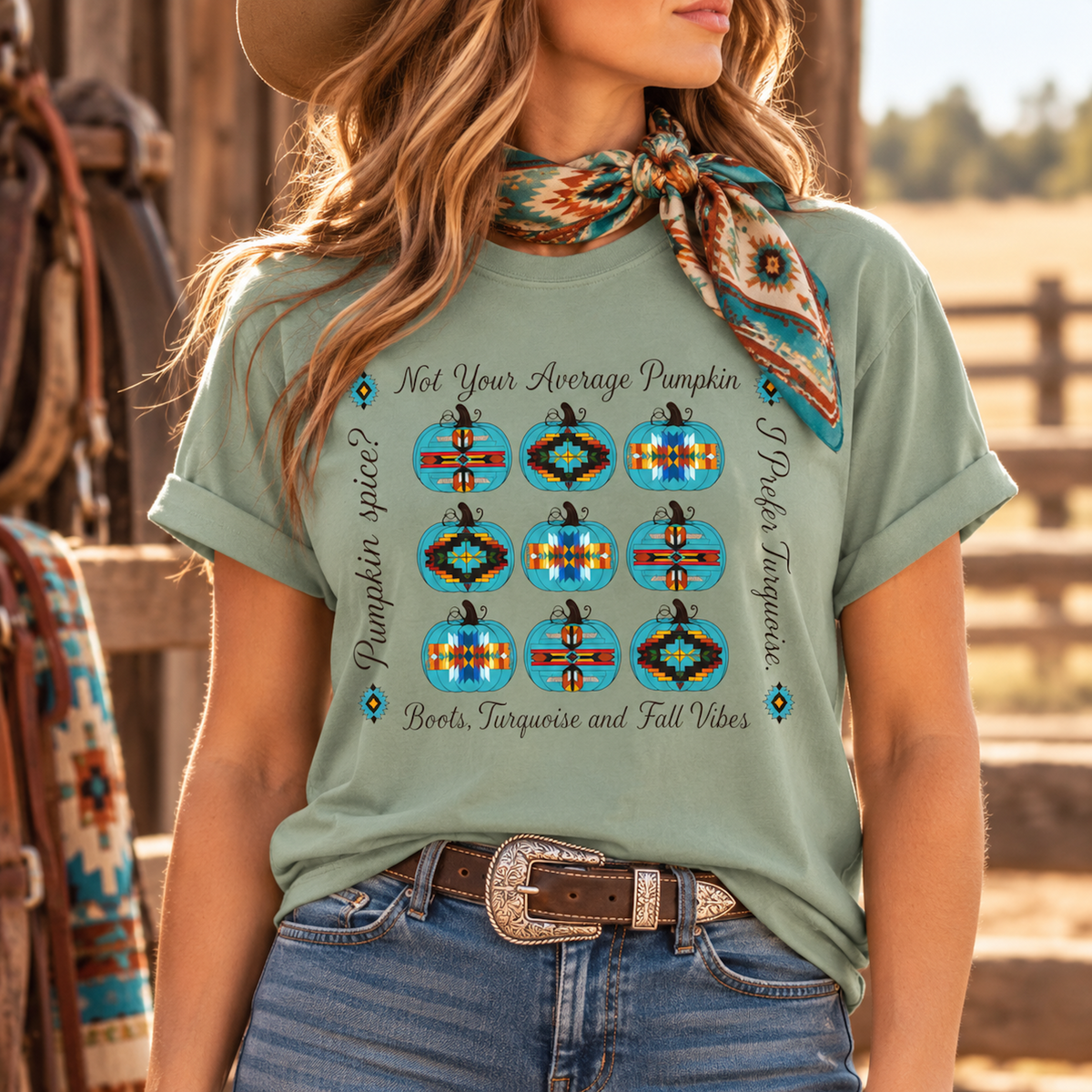 A woman wearing a sage green graphic t-shirt with a southwestern-themed pumpkin print, styled with a matching patterned neck scarf and jeans in a rustic outdoor setting.