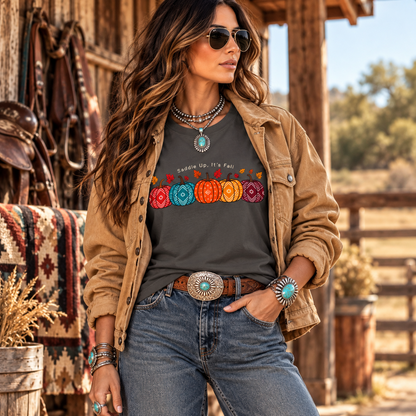 A woman wearing a dark gray graphic t-shirt with a colorful pumpkin design and the text 'Sudden Up, It's Fall', styled with a tan jacket, blue jeans, and western accessories in a rustic outdoor setting.