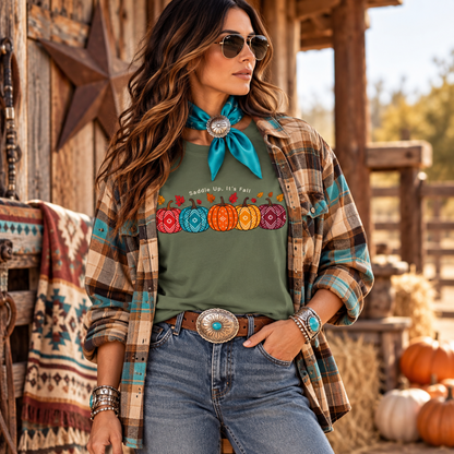 A woman stands outdoors in a rustic barn-like setting wearing a sage green graphic T-shirt with a colorful embroidered pumpkin row and the words 'saddle up It's Fall,' layered under an open plaid flannel shirt with a turquoise neck scarf and blue jeans.