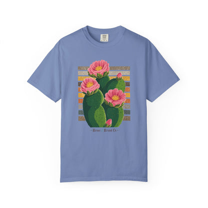 A light blue crewneck t-shirt featuring a graphic print of a green prickly pear cactus with pink flowers and yellow centers, set against a striped background with the text '-Brass Brand Co.-' printed below.