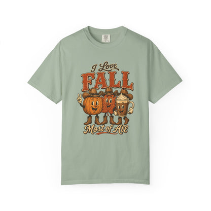 A sage green short-sleeve t-shirt with a vintage-style autumn graphic on the chest. The graphic features the text 'I Love FALL Most of All' in brown and orange lettering, accompanied by three cartoon characters dressed as pumpkins, a gingerbread man, and a mug of coffee.