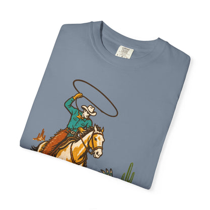 A folded slate blue crewneck t-shirt featuring a western graphic of a cowboy on a horse swinging a lasso, displayed on a clean white background.
