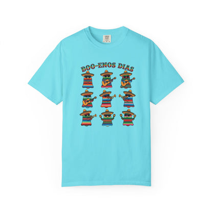 A light blue short-sleeve t-shirt with a colorful graphic print on the front. The graphic features the text 'BOO-KENOS DIAS' above nine cartoon characters wearing sombrero hats and colorful striped ponchos, each holding a guitar or drum, set against a plain white background.