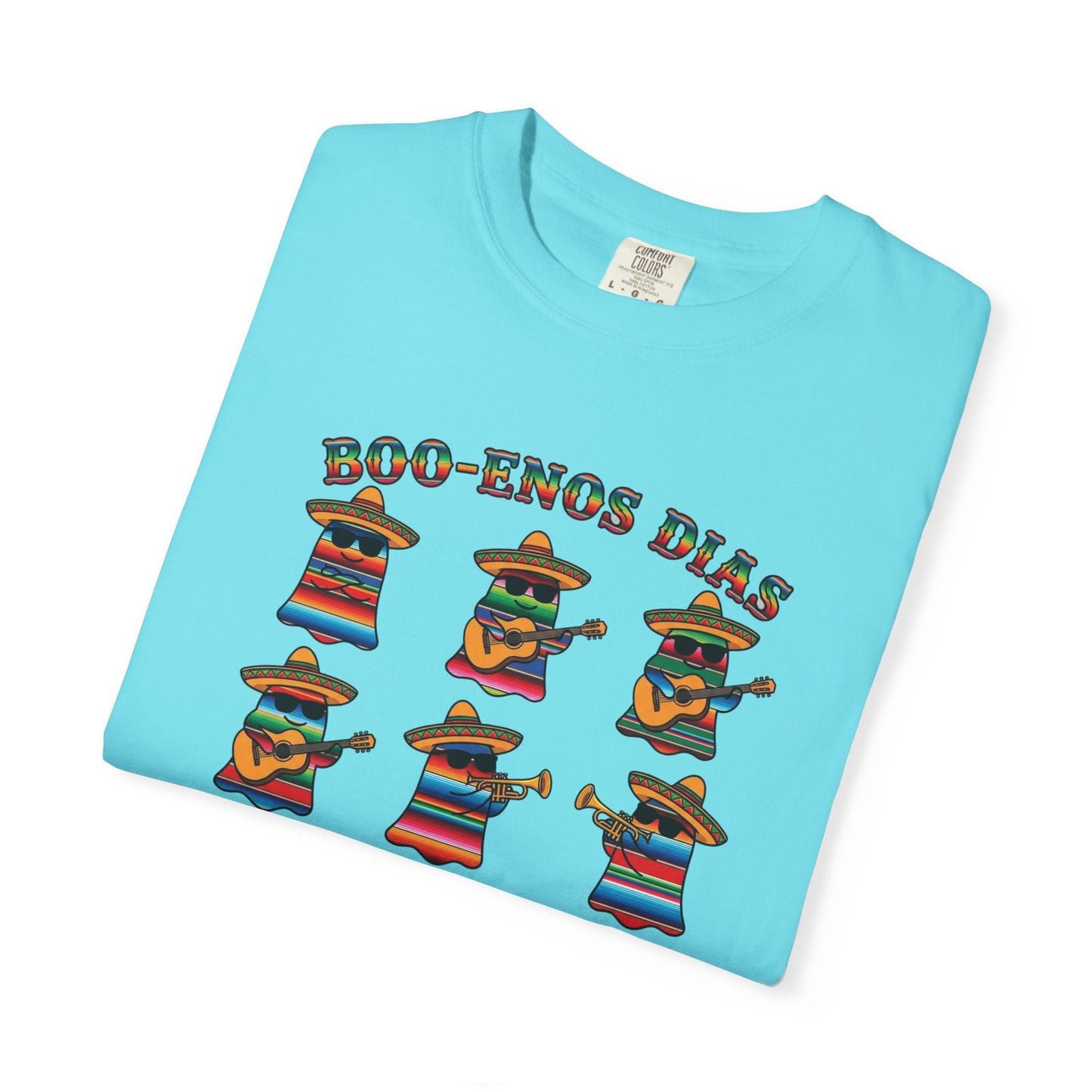 A folded light blue t-shirt on a white background, featuring a colorful graphic of six cartoon characters dressed in Mexican serape patterns and sombreros, playing musical instruments, with the text 'BOO-ENOS DIAS' above them.