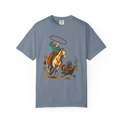 A slate blue crewneck t-shirt featuring a colorful graphic print of a cowboy riding a horse and lassoing a turkey, displayed on a clean white background.