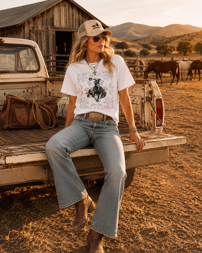 Woman sitting on a pickup truck bed in a rural setting with mountains in the background in white paisley rodeo tee